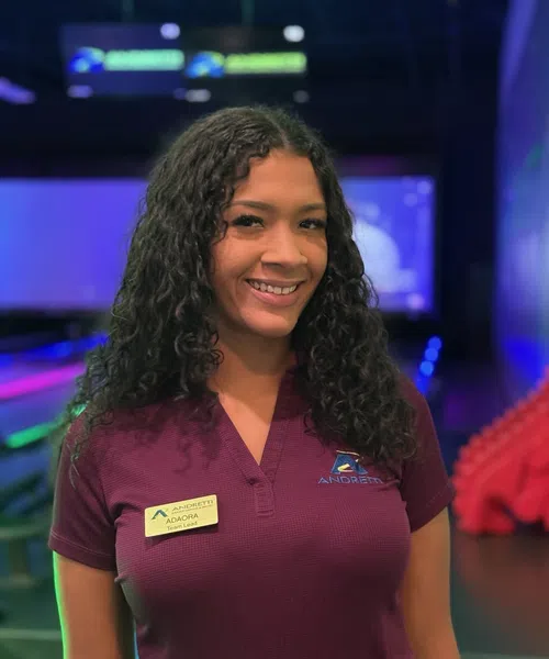 Smiling woman with curly hair wearing a maroon Andretti team shirt and badge standing in an indoor arcade or gaming center.