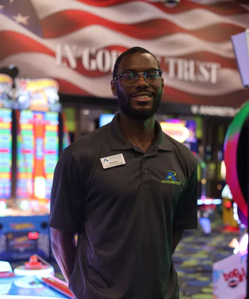 Smile assistant wearing Andretti uniform stands in front of arcade games with a blurred American flag backdrop.