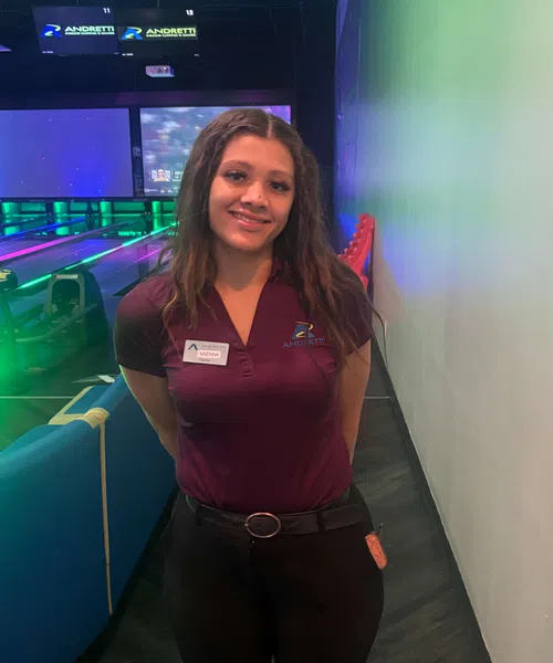 Smiling Andretti Indoor Karting staff member in maroon shirt stands near bowling lanes with neon lights.