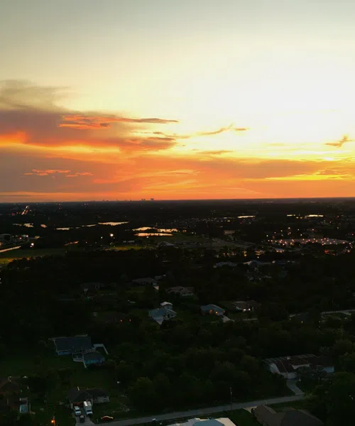 Aerial view of a suburban neighborhood at sunset with colorful sky and scattered houses surrounded by trees.