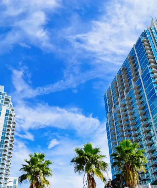 Modern glass skyscrapers with balconies and palm trees under a bright blue sky with wispy clouds