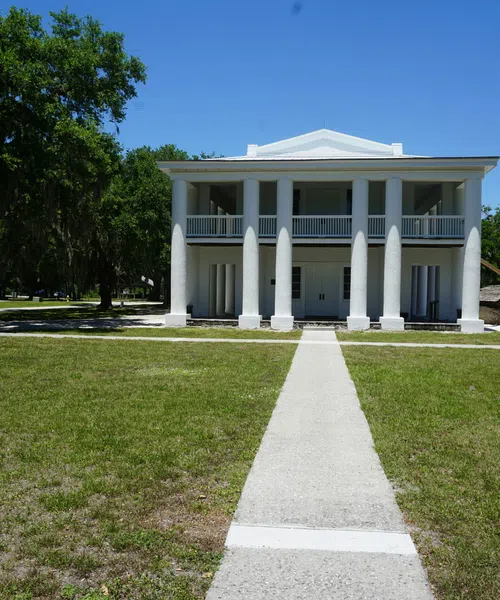 White historic two-story building with columns, surrounded by green lawn and trees under clear blue sky.