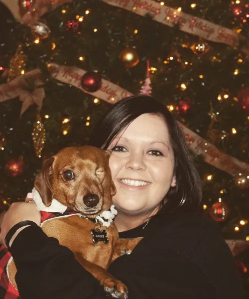 a smiling woman holding a one-eyed dog in front of a Christmas tree