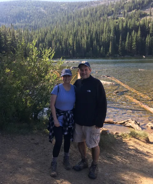 a man and a woman standing before a lake in a wooded area