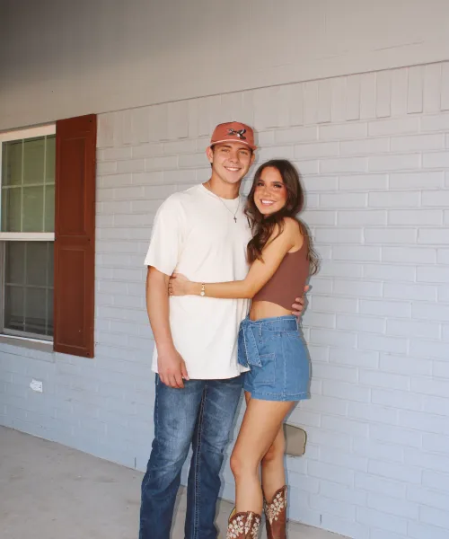 Couple posing happily in casual outfits with cowboy boots, standing against a light brick wall.
