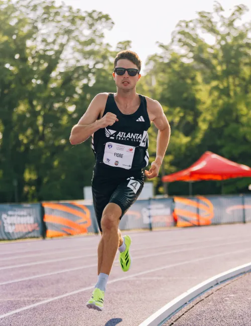 Male athlete in black running gear sprints on outdoor track during sunny day with trees and tents in the background