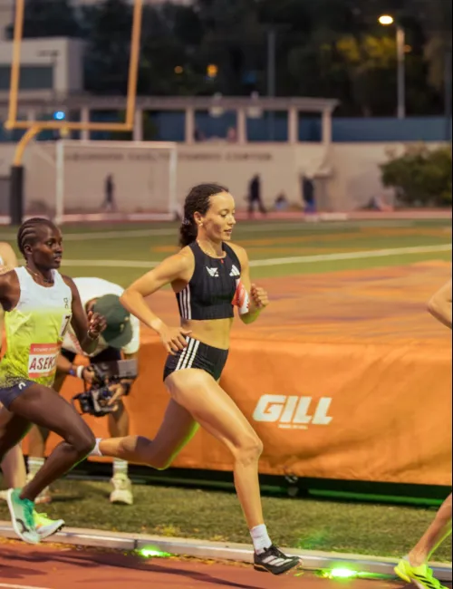 Female athletes compete in a middle-distance race on an outdoor track during evening.