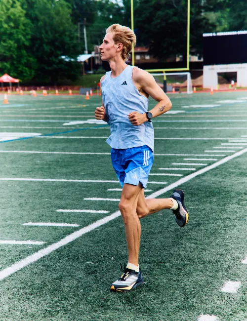 Athlete running on a football field wearing blue shorts, tank top, and sport shoes during daytime training.