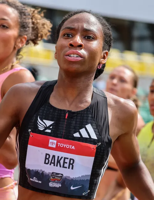 Female athletes running in a competitive outdoor track race, focusing on runner with bib labeled Baker.