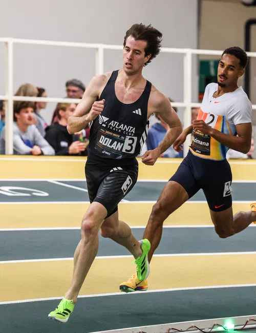Two male athletes sprinting in an indoor track race with spectators watching from the stands
