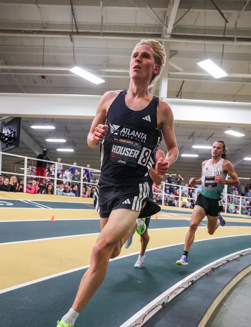 Indoor track runner wearing black Atlanta Track Club uniform leads a race on curved track with competitor behind.