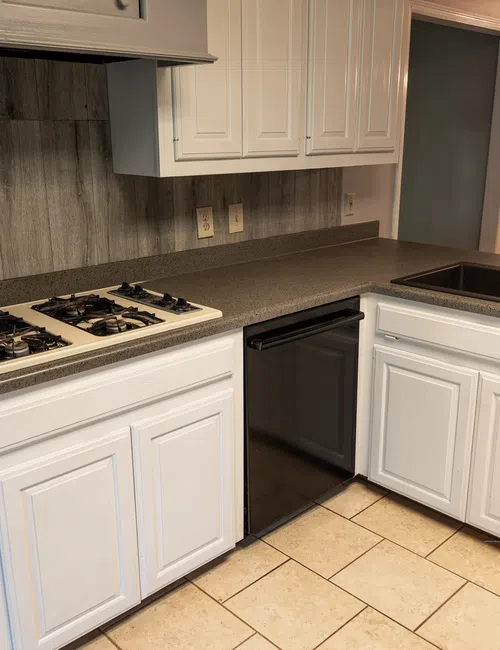 Modern kitchen with white cabinets, black sink, gas stove, and beige tiled floor under natural light.