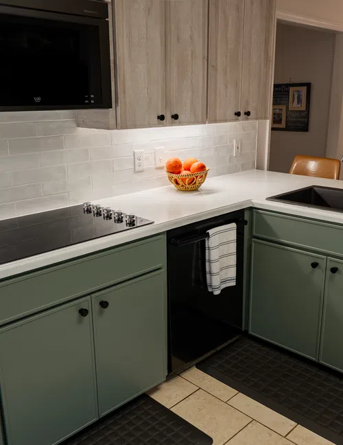 Modern kitchen with green cabinets, white countertops, black sink, stovetop, and utensil holder under warm lighting
