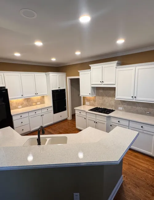 Modern kitchen with white cabinets, quartz countertops, hardwood floors, and built-in black appliances under recessed lighting.