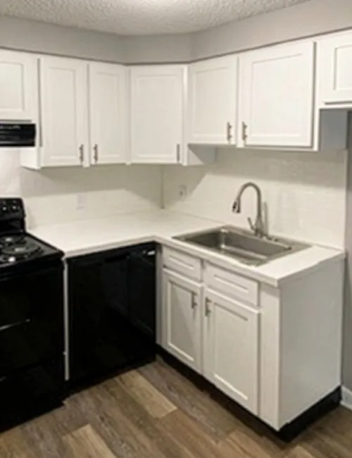 Modern kitchen with white cabinets, black appliances, a stainless steel sink, and wood flooring under bright ceiling light.