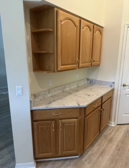 Kitchen cabinetry with oak wood finish and marble-patterned countertop against beige walls and light hardwood floor.