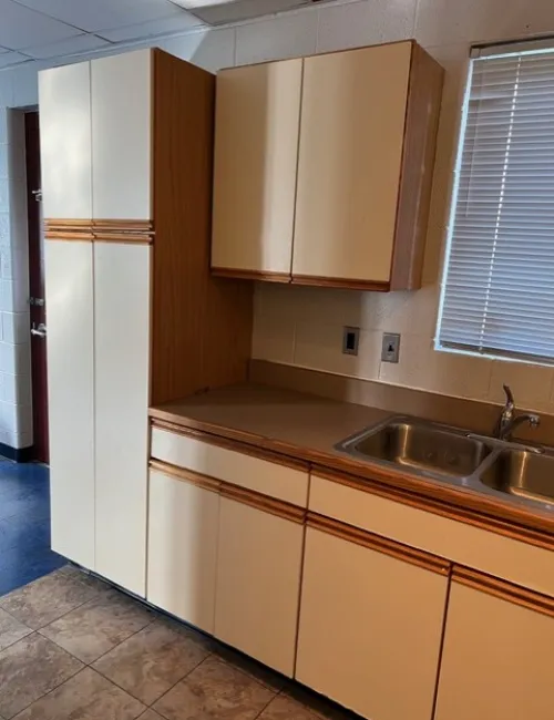 Compact kitchen area with cream cabinets, double stainless steel sink, tiled floor, and window with blinds.