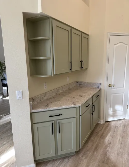 Light green kitchen cabinets with black handles and beige marble countertop in a corner space by a white door.
