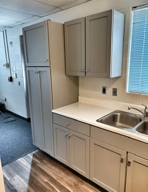 Modern kitchen area with gray cabinets, white countertop, double stainless steel sink, and window with blinds.