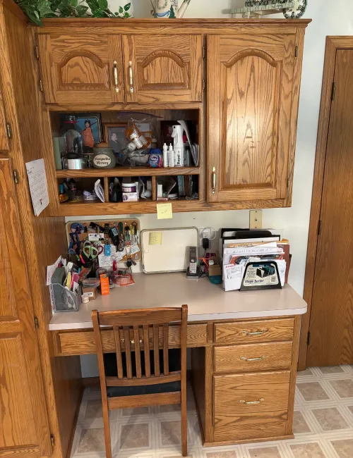 Cluttered wooden desk with papers, keys, medication, and office supplies next to a wooden chair and cabinets.