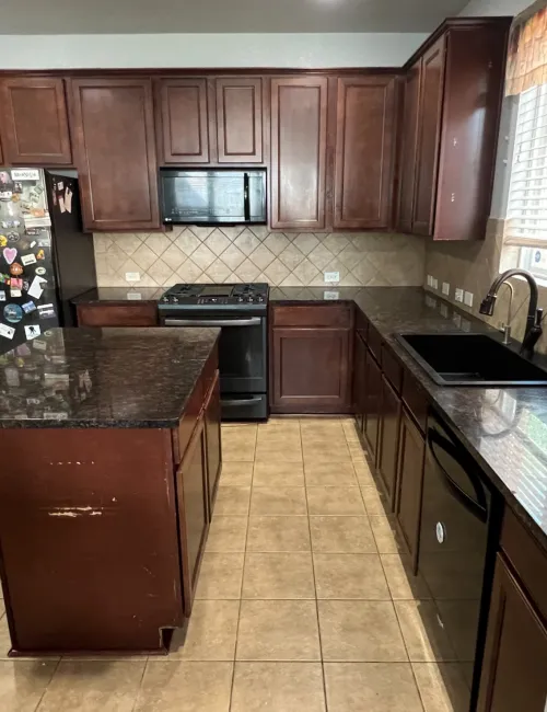 Modern kitchen with dark wood cabinets, black appliances, granite island, and tiled flooring in natural light.