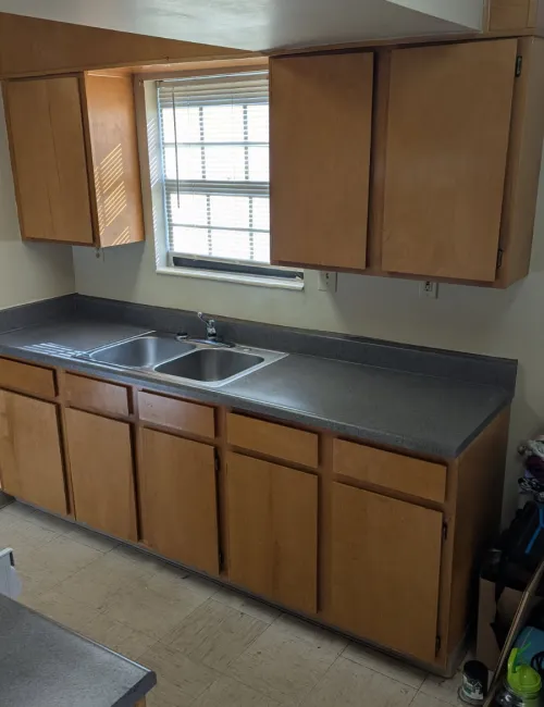 Compact kitchen with wooden cabinets, double stainless steel sink, gray countertop, and window with blinds.