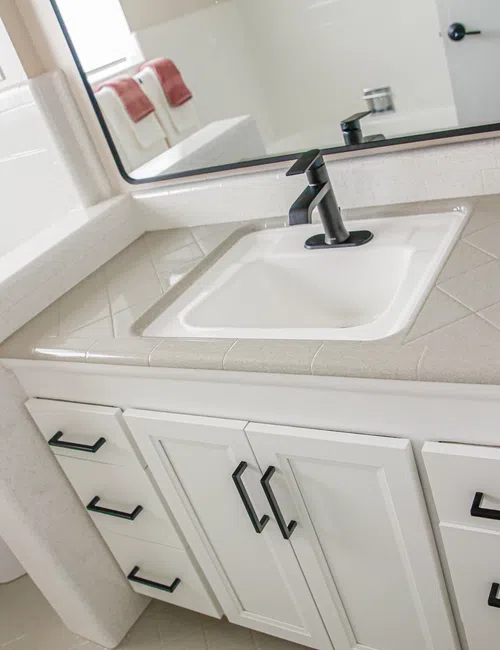 Modern bathroom sink with white cabinetry and black faucet, beige tiled countertop, and red towels in the background
