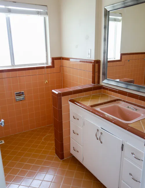 Vintage bathroom with orange tiled walls and floor, pink sink, white cabinets, and a large mirror.