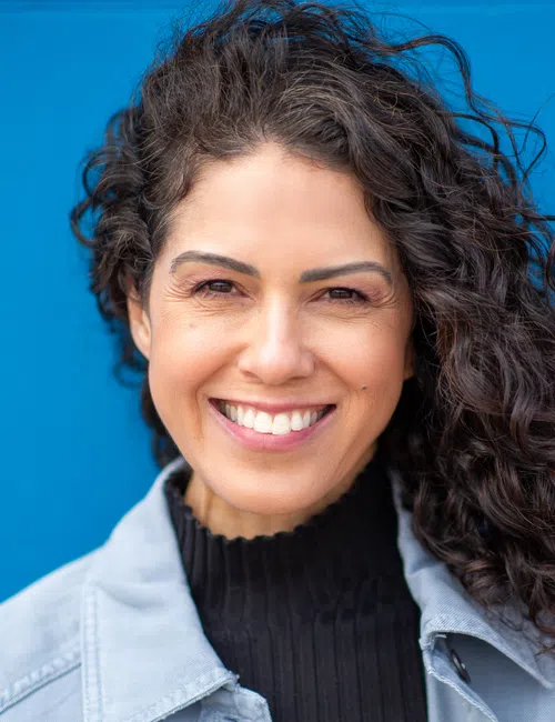 Smiling woman with curly dark hair wearing a light jacket and black top against a blue background