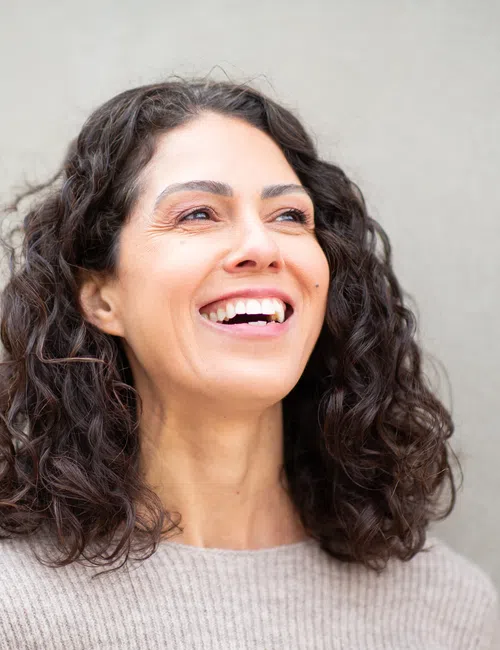 Happy woman with curly dark hair smiling against a plain beige wall background.