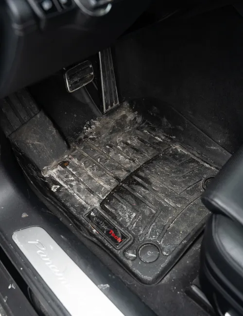 Dirty black car floor mat with mud and dust inside a vehicle near the brake pedal and door sill.