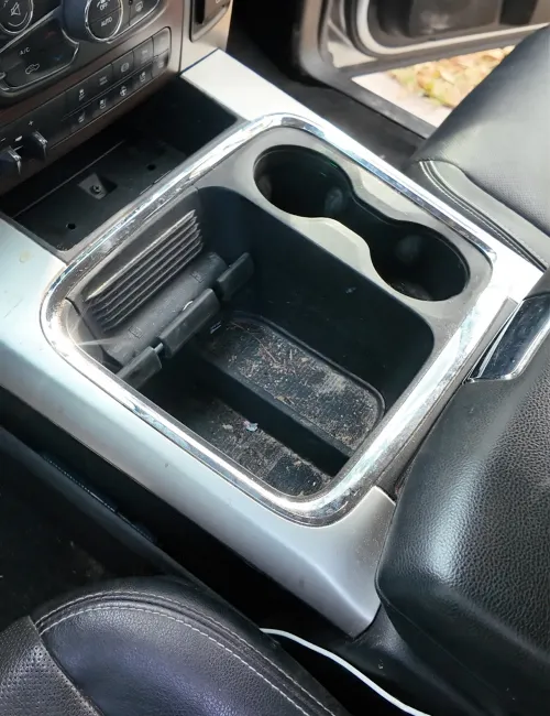 Dirty car center console with two cup holders and a storage compartment between black leather seats.