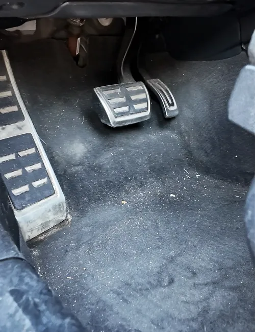 Dirty car floor mat with brake and accelerator pedals in a vehicle's driver's side footwell.