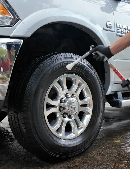 Close-up of a person cleaning a silver RAM 2500 truck tire with a foam spray applicator.