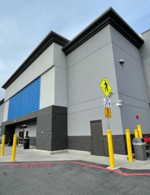 Modern commercial building with gray and blue exterior, yellow safety poles, and pedestrian crossing signs in parking lot.