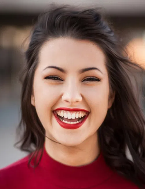 Young woman in a red top smiling brightly with wavy hair outdoors in soft lighting.