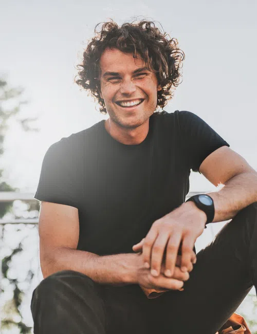 Smiling young man with curly hair wearing black t-shirt sitting outdoors with trees and railing in background