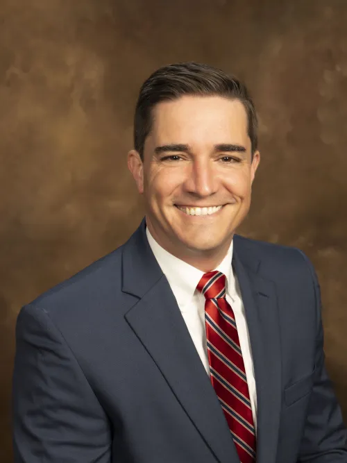 Smiling man in a suit with a red tie, posing against a warm, textured background.