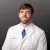 Smiling male doctor in white coat and blue tie against gray background, representing advanced urology care.
