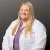 Smiling female medical professional with long blonde hair wearing a white lab coat and purple top on gray background
