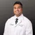 Smiling male doctor in white coat and green tie posing against dark gray background in professional headshot.