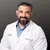 Smiling male doctor with beard wearing white lab coat and blue shirt against a neutral gray background