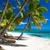 Tropical beach with white sand, leaning palm trees, wooden lounge chairs, and clear turquoise ocean under blue sky.