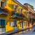 Colorful yellow buildings with flower-filled balconies lining a sunny street with a blue tuk-tuk parked nearby.