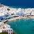 Aerial view of Mykonos town with white buildings, blue sea, boats, and windmills under a clear sky.