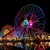Night view of a brightly lit Mickey Mouse Ferris wheel and roller coaster with colorful reflections on water.