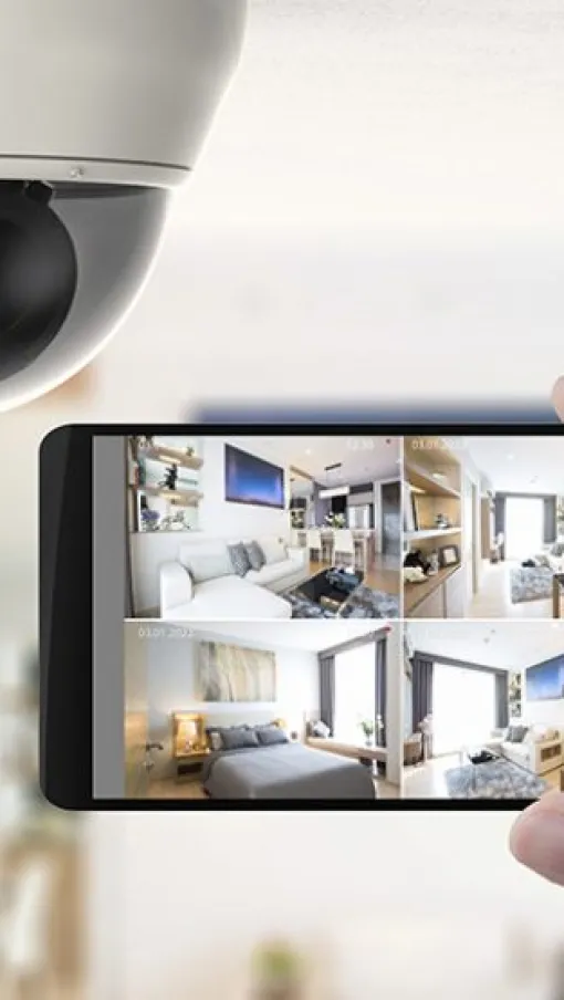 Hand holding smartphone displaying home security camera footage under white surveillance dome camera indoors.