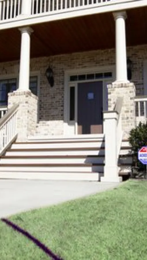 Front porch of a brick house with white columns, staircase, and a security system sign on the lawn.