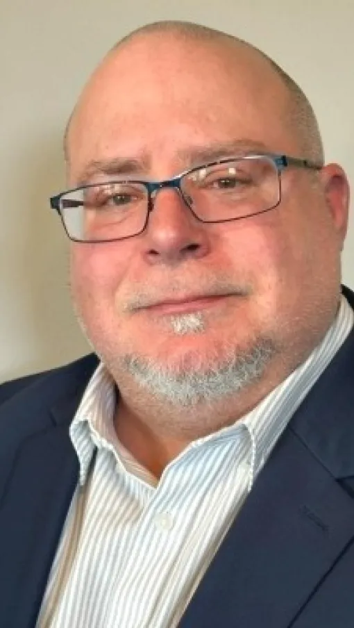 Middle-aged man with glasses, bald head, and gray beard wearing a navy blazer and striped shirt against a neutral background