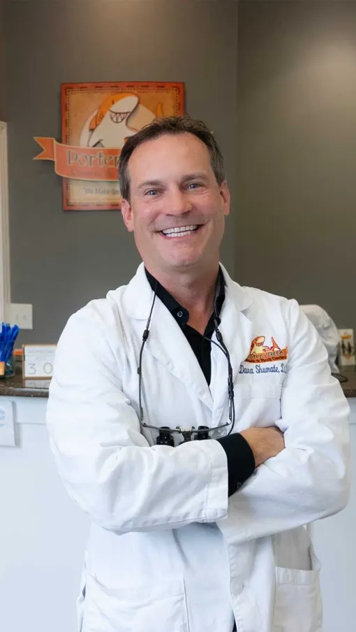 Smiling male dentist in white coat with crossed arms standing in modern dental office reception area.